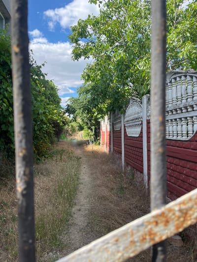 The image depicts a narrow dirt path bordered by dry grass and lush green foliage, leading into the distance under a partly cloudy sky. On the right side of the path, a red and white decorative fence runs along its length, with a portion of a large green tree visible above it. The foreground is dominated by blurred vertical metal bars and a rusty horizontal bar, suggesting the photo was taken through a gate or fence. The overall scene suggests an outdoor location, possibly a pathway in a residential or rural area in Kryzhanivka, Ukraine. No people or specific actions are visible.