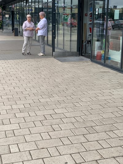 Two elderly women are standing on a paved walkway in front of a store with large glass windows. The woman on the left is wearing a light purple shirt and light gray pants. She has short gray hair and glasses, and is looking at the woman on the right. The woman on the right is wearing a white patterned shirt and gray pants. She has short gray hair and is looking at the woman on the left. The store appears to be a retail establishment. The walkway is made of light-colored rectangular paving stones. The background shows reflections in the store windows, including cars and other buildings. The scene suggests a casual encounter or conversation outdoors.