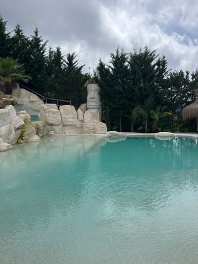 The image shows a swimming pool with clear turquoise water and a sandy bottom. The pool is surrounded by artificial rock formations and lush green trees, including palm trees and cypress trees. A large tiki statue is visible behind the rock formations. In the background, a thatched-roof hut can be seen. The sky is overcast with clouds. The location is identified as Can Güell, Spain.