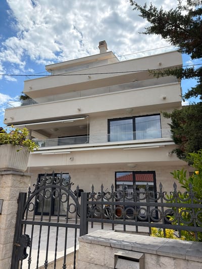A modern building with staggered balconies and large windows is visible behind a dark metal fence. The building's facade is light-colored, possibly stone or concrete. Some windows reflect the sky, indicating they are closed. The fence has decorative circular and spear-shaped elements. Above the building, there's a partial view of trees and a blue sky with clouds. A mailbox with the word "POSTA" is near the fence on the right. The location appears to be Krašići, Montenegro, judging by the style of the building and fence. There are no people visible in the image.