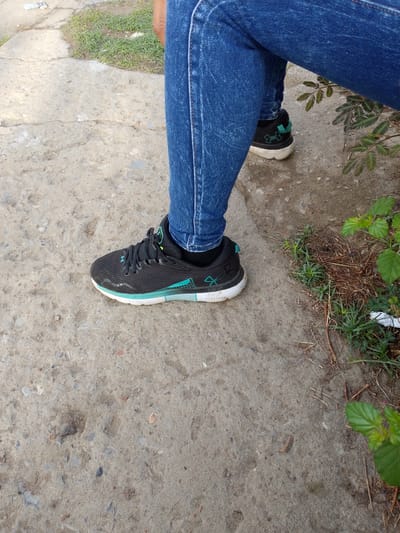 A person wearing blue jeans and black Under Armour HOVR sneakers is sitting on a concrete surface. The concrete surface shows signs of wear and tear. There is vegetation present, including grass and leafy plants. The specific location within Barcelona, Venezuela, is not discernible from the image alone.
