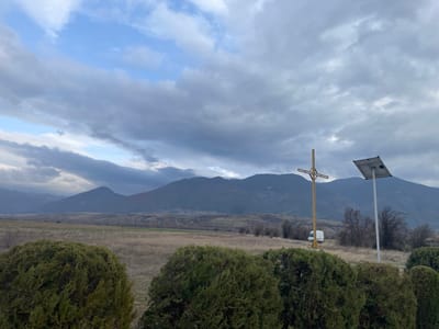 The image depicts a rural or semi-rural landscape in Krainitsi, Bulgaria, under an overcast sky. A range of dark, undulating mountains forms the background, partially obscured by heavy grey clouds with intermittent patches of lighter blue sky. The midground consists of expansive, dry, light brown fields, indicative of late autumn or winter. In the foreground, three dense, dark green evergreen shrubs partially frame the scene. Centrally positioned behind these shrubs, a tall, light-colored wooden or metal Christian cross stands upright. To the right of the cross, a white pole supports a rectangular solar panel angled towards the sky. Further in the midground, beyond the cross and pole, a white van is visible on what appears to be a dirt road or track. No individuals are present.