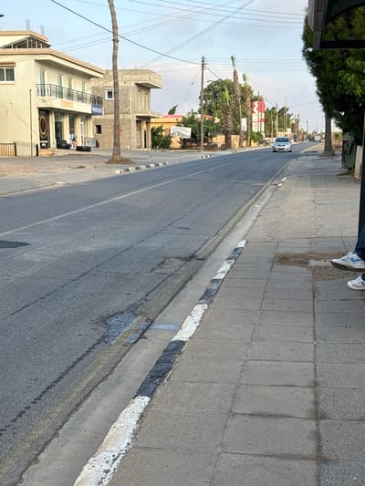 The image captures a street scene in Xylofagou, Sovereign Base Areas of Akrotiri and Dhekelia. A light-colored car is visible on the road, heading towards the camera. On the right side of the road, a person is seated on a sidewalk, their legs and feet visible. They are wearing blue jeans and white sneakers. Adjacent to the sidewalk is a paved road with painted white and black curb markings. Buildings line the left side of the street, including a light-colored, multi-story structure with balconies and shops on the ground floor. Palm trees and other greenery are interspersed along the street. Overhead, power lines crisscross the sky. The overall scene depicts a quiet, daytime street environment.