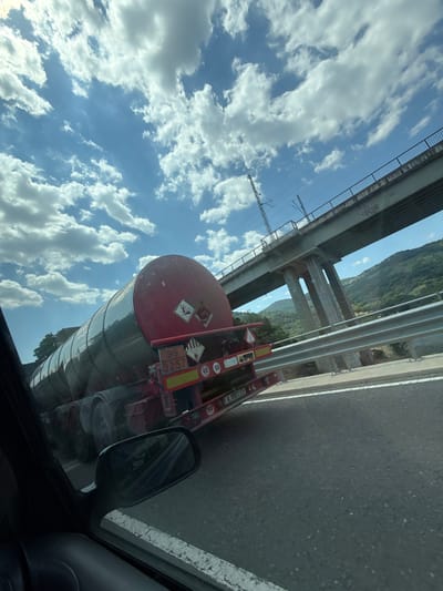 The image captures a view from a vehicle on a highway, looking towards the right side. A large red tanker truck is prominently featured, traveling on the same highway. The truck displays hazard symbols indicating flammable liquids and toxic substances. It also has a license plate that appears to read "A 8882 EE".
Above the highway, a concrete overpass for a railway line stretches across the scene, supported by multiple pillars. The sky is a vibrant blue, dotted with fluffy white clouds, suggesting fair weather. In the background, a lush green, mountainous landscape is visible.
The perspective suggests the photo was taken while moving, indicated by the slight blur and the angle of the shot. The interior of the vehicle from which the photo was taken is partially visible on the left, including a side mirror and part of the car's interior. The overall environment points to a location on a highway in a semi-rural or mountainous area, possibly near Asparuhovo, Bulgaria, given the context.