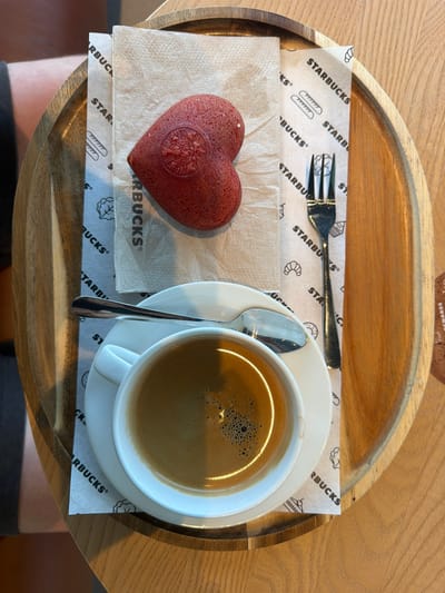 The image shows a top-down view of a wooden tray holding a Starbucks order. On the left, there is a heart-shaped red velvet pastry resting on a white napkin. To the right of the pastry is a white cup of coffee on a white saucer, accompanied by a silver spoon. A silver fork rests near the top of the tray. All items are placed on a white paper mat printed with the Starbucks logo and various pastry illustrations. The tray itself has a curved, raised edge. The tray is sitting on a wooden table, with a portion of someone's lap visible in the lower part of the image. There is no indication of the location in Weil am Rhein, Germany.
