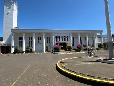 In the city of Desa Tambakreja, Indonesia, a modern white building with a prominent tower stands under a clear blue sky. The building features a colonnaded facade with a prominent "KA Cilacap" logo above the entrance. In front of the building, a paved area serves as a parking lot and a circular driveway. A lone bicyclist is captured in motion on the driveway, casting a shadow on the asphalt. Orange traffic cones and neatly trimmed bushes adorn the area, alongside a "VIP" sign near the entrance. The scene is bathed in bright sunlight, indicating a warm, clear day.