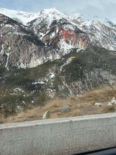This image shows a mountain landscape in Montgenèvre, France. The scene is captured from an elevated position, likely from a road or structure with a concrete barrier visible in the foreground. The mountain is partially covered with snow and features rocky terrain, including areas with red soil. Patches of trees are interspersed throughout the mountainside. The sky is overcast. Dry grass grows near the concrete barrier.
