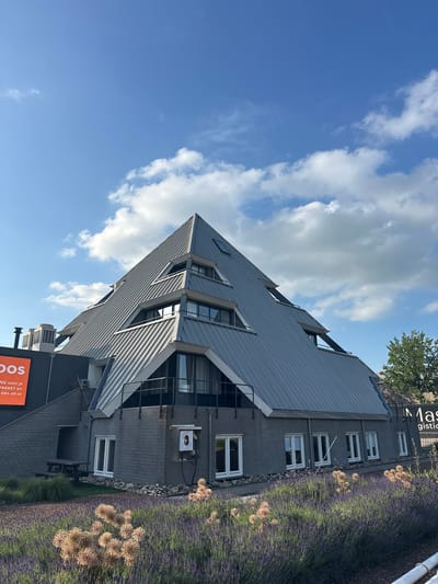 The image shows a modern, pyramid-shaped building with a grey metal roof, located in the city of Staphorst, Netherlands. The building has multiple levels with balconies and several windows. In the foreground, there is a garden with lavender and dried ornamental plants. To the left of the building, an orange sign with Dutch text is visible. A tree is present to the right of the building. The sky is blue with scattered white clouds. The building appears to be a commercial property, possibly an office or business.