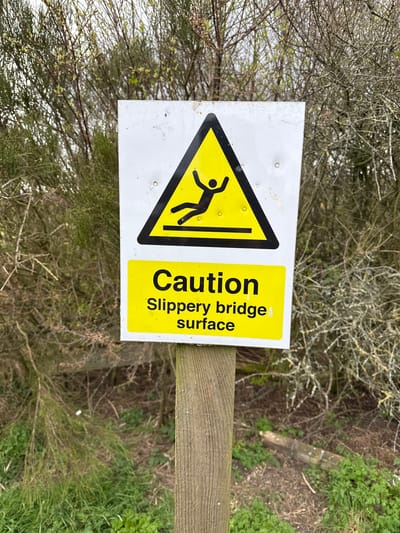 A rectangular sign, mounted on a wooden post, reads "Caution Slippery bridge surface". The sign features a yellow triangle with a black silhouette depicting a falling figure. The backdrop consists of dense foliage, suggestive of a natural setting. The location is presumed to be in Dunfermline, United Kingdom.