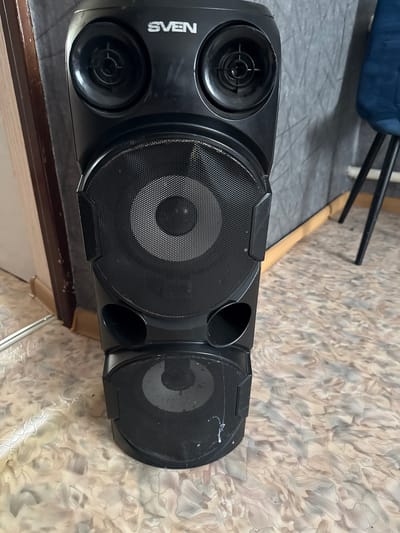 A black Sven brand speaker tower stands on a patterned floor in a room. The tower has two large circular speaker grilles on the front, with smaller circular vents located between them. Above the main speakers are two smaller, raised circular speaker cones. The top of the speaker has the "SVEN" logo in white text. To the left of the speaker is a wooden door frame, and to the right, a dark blue chair is visible. The wall behind the speaker is a textured grey. The floor is covered in a light-colored linoleum with an abstract pattern. The location is specified as Айет, Kazakhstan.