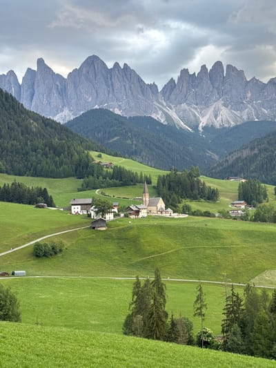 The image depicts the village of St. Magdalena in the Val di Funes, South Tyrol, Italy, with the iconic Odle/Geisler peaks forming a dramatic mountain backdrop. The scene is characterized by rolling green hills dotted with traditional Tyrolean farmhouses and chalets. A prominent church with a tall spire stands in the center of the village. Verdant meadows, characterized by their lush green grass, dominate the foreground and midground, with winding pathways and roads cutting through them. Scattered trees and small clusters of pine forests add texture and depth to the landscape. The sky above is overcast with soft, diffused light, contributing to a serene and picturesque atmosphere. A few indistinct figures can be seen walking along a path in the midground. The overall impression is one of a peaceful, idyllic alpine village nestled within a majestic mountain range.