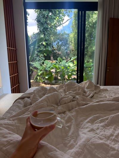 Here's a detailed description of the image:
**Scene Overview:** The image captures an indoor scene, possibly a bedroom, with a view to the outside. A hand holds a glass cup with a light-colored liquid (likely coffee or tea) in the foreground. A rumpled, light-colored bedspread is also visible.
**Environment:** A large window frames a view of lush greenery. The vegetation outside appears dense and tropical, possibly a garden or forest. The presence of these plants suggests a tropical environment. The wooden blinds are visible under the window.
**Objects:**
* **Glass Cup:** Contains a light-brown liquid (coffee or tea). The cup has a handle.
* **Bedspread:** Light-colored and rumpled, resting on a bed. The bedspread appears to have a quilted pattern.
* **Window:** Large, with a black frame, offering a view to the outside.
* **Vegetation:** Dense greenery is visible outside the window.
**Action:** A hand is holding a glass cup of coffee over the bed.
**People:** One hand is visible.
**Location:** Mas, Indonesia is likely based on the tropical greenery and overall vibe of the image.