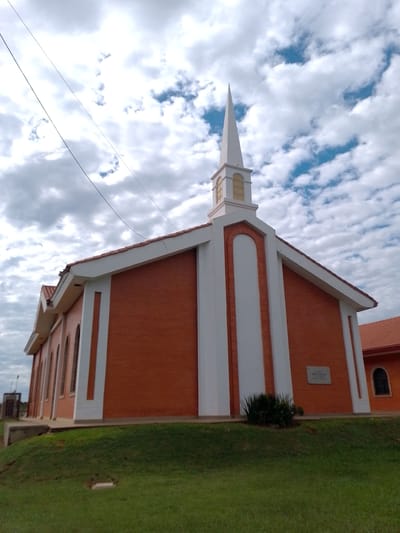 The image captures a Latter-day Saint church building in Concepción, Paraguay. The building is constructed of red brick with white trim. It features a prominent white steeple. The building is set on a grassy lawn under a cloudy sky. Overhead power lines are visible. A sign, likely bearing the name of the church, is affixed to the right side of the building. Another, smaller, building is seen on the right.