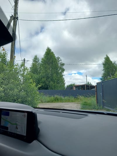 The image is taken from inside a car, looking forward. The dashboard of the car is visible in the foreground, with a navigation screen displaying a map on the left side. Outside the car, there is a dirt road leading to a dark gray metal gate that is partially open. Beyond the road, there is a dark gray fence. Behind the fence, there are trees and a building with a wooden roof structure. Overhead, there are power lines and a cloudy sky with some patches of blue. The overall scene depicts an outdoor environment, possibly a rural area or the outskirts of a town. The location is identified as Verkhneye Dubrovo, Russia. There are no people visible in the image.