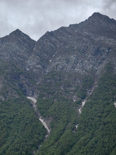 The image shows a mountain range in the city of Gondhla, India, under a cloudy sky. The lower slopes are covered in dense green trees, while the upper, steeper sections are rocky and barren, with patches of snow visible. The mountain peaks are sharp and jagged. There are no people or animals visible in the image.