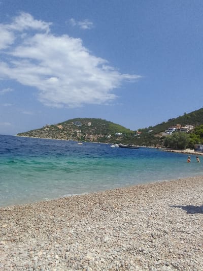 The image depicts a tranquil beach scene on a sunny day. A wide expanse of pebbled shore stretches across the foreground, meeting the clear, turquoise waters of the sea. The water gradually deepens into a rich blue further out, where several boats are visible.
In the middle ground, a hilly coastline is dotted with white buildings, suggesting a small settlement or village. The hills are covered with sparse vegetation. The sky is a vibrant blue, with scattered white clouds.
In the right middle ground of the water, two people are visible, appearing to be swimming or wading. Their presence adds a sense of activity to the otherwise serene landscape. The overall impression is one of a beautiful and relaxing coastal environment, likely within the Municipal Unit of Solygeia, Greece, known for its scenic beaches and villages.