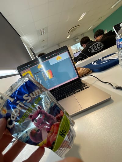 The image depicts an indoor scene within a classroom or computer lab setting, identified as likely a Tremplin Numérique facility in Bron, France.
In the immediate foreground, a left hand holds a crumpled Capri-Sun "Monster Alarm" juice pouch. The pouch is predominantly blue and silver, featuring a pink monster character and illustrations of fruits, with a white straw inserted. Text indicating "NO ARTIFICIAL PRESERVATIVES COLOURS" is visible.
Centered on a white, textured desk is an open silver MacBook Pro laptop. Its screen displays a webpage from `tremplinnumerique.org`, featuring the "Tremplin Numérique" logo, a search bar labeled "Trouver un mot sur Wikipédia," and a data table with numerical percentages (e.g., 89.04%, 75.00%) alongside horizontal colored gradient bars. A black power cable is connected to the right side of the laptop.
To the right of the laptop, on the same desk, are a partially full 1.5-liter clear plastic bottle of Cristaline mineral water and a blue zippered pouch with white wired earbuds resting on top. A crumpled brown paper bag is situated between the laptop and these items.
In the background, three individuals are seated at similar white desks. One individual, most prominently visible, wears a black hoodie with a white wave-like graphic on the back and a black beanie, facing away from the camera towards a screen. The room features a white grid drop ceiling with recessed rectangular fluorescent light fixtures and square air vents. A large screen or window in the far background displays an outdoor scene with bare trees under natural light.