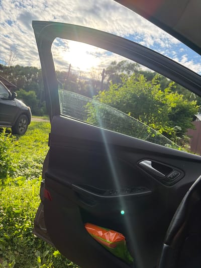The image captures a view from inside a car, looking out through the open driver's side door. The door panel is visible in the foreground, featuring a silver door handle and buttons for window controls and door locks. A bright orange package, possibly containing food or supplies, is tucked into a compartment on the door. Sunlight streams in from the left, creating lens flare and illuminating the car's interior.
Through the open door and window, a sun-drenched outdoor scene unfolds. The sky is a vibrant blue with scattered white clouds. Lush green trees and foliage dominate the background, suggesting a natural or semi-rural setting. The sun appears to be positioned low in the sky, casting long shadows and indicating either early morning or late afternoon. The overall impression is one of a bright, sunny day, likely in a temperate climate.
In the lower portion of the image, the rear wheel and part of the exterior of another vehicle are visible. This suggests the car the photo is taken from is parked, possibly on a grassy or unpaved area, as indicated by the green vegetation and the presence of small white flowers on the ground. The light quality and the surrounding greenery suggest a warm season, likely summer. No people are visible in the image. The image appears to be taken in Kimry, Russia.