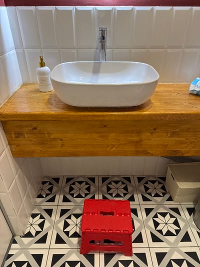 This media file captures a detail of a bathroom or lavatory space in a home in Patrusheva, Russia. The scene features a white urinal mounted on a wooden column. The wall behind the urinal is tiled with white subway tiles. To the right of the urinal, there is a section of floor tiled with a black and white geometric pattern, and a red foldable stool is visible. A white soap dispenser with a gold pump is attached to the wall near the base of the urinal. The lighting suggests it is daytime, likely indoors. No people are visible. There is some text on the soap dispenser, "Ditta ssa", and on a visible item at the top of the image, "Tork".