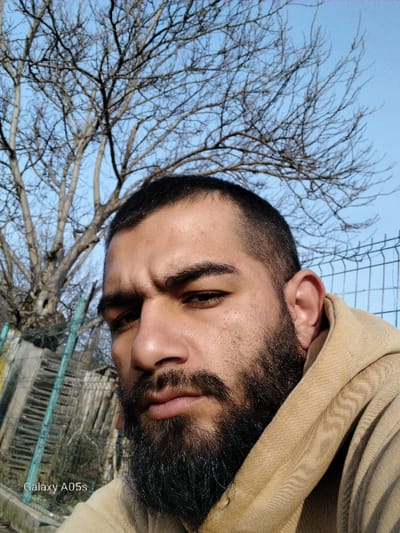 A close-up, upward-angled shot depicts a male individual outdoors in Oreshene, Bulgaria. The subject, positioned in the lower two-thirds of the frame, has dark hair with a faded haircut on the sides and a prominent, full dark beard. He wears a light brown or tan hooded sweatshirt, with the hood visible on his right side. His gaze is directed towards the camera with a neutral expression. The background features a clear blue sky occupying the upper third of the image. A large, bare deciduous tree with extensive branching is centrally visible against the sky. To the left of the subject, a dilapidated wooden or corrugated metal fence structure is present, along with what appears to be part of a chain-link fence. To the right, a chain-link fence is also visible. The ambient light suggests daylight. A "Galaxy A05s" watermark is present in the lower-left corner of the image.