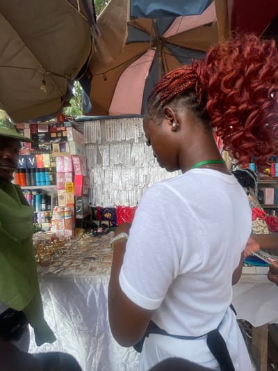 This media file depicts a vibrant market scene in Ise- Ekiti, Nigeria, likely during daytime given the bright natural light. The main subject is a woman with striking red braided hair, wearing a white t-shirt and a green necklace. She is viewed from the side, her head tilted downwards as she appears to be engaged with the market stall. Her expression is focused and perhaps a little contemplative.
The market stall itself is a riot of colors and products, suggesting a bustling commercial environment. Various items are displayed, including an assortment of jewelry such as necklaces, bracelets, and earrings, presented in clear packaging and on display racks. There are also numerous boxed products, some resembling cosmetics or personal care items, with visible branding. Among these are bottles of what appear to be deodorants or body sprays. The stall is partially covered by umbrellas, indicating an outdoor setting where protection from sun or rain is necessary.
Other individuals are present, though less prominently featured. One person, wearing a green cap, is visible in the lower portion of the image, also seemingly involved in the market activities. The overall atmosphere is one of lively commerce, with a focus on everyday goods and accessories. The presence of diverse products and the organized display suggest a well-established marketplace catering to local needs. The image captures a moment of interaction within this dynamic environment.