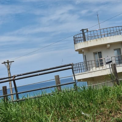 A white, multi-story building with a balcony railing, and a lightning rod on top, stands against a blue sky with scattered clouds. A metal fence with a rope railing runs in front of the building, with tall green grass in the foreground. The ocean is visible behind the fence. A sign in Korean is attached to the building's exterior. The scene appears to be in Gangneung-si, South Korea, based on the Korean text on the sign.