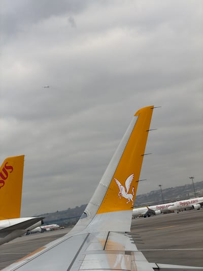 A forensic examination of the image reveals a partial view of an airport tarmac scene, presumably at Sabiha Gökçen International Airport, located near Tuzla, Istanbul, Turkey.

In the foreground, the right wing and yellow vertical stabilizer of a white Pegasus Airlines aircraft are prominently displayed, viewed from an interior window. The vertical stabilizer features a white Pegasus logo and the airline's name, "PEGASUS," is partially visible on an adjacent tail fin to the left. The aircraft's wing surface shows minor dirt accumulation.

In the mid-ground, several other white Pegasus Airlines aircraft, identifiable by their yellow tail fins and "flypgs.com" branding on their fuselages, are parked on an asphalt tarmac. Yellow line markings are visible on the tarmac surface.

The background features a distant, rolling green hill, topped with multiple tall light poles and indistinct urban or industrial structures. The sky above is uniformly overcast with a dense layer of light grey clouds. A small, white aircraft is discernible in the upper left quadrant of the sky, appearing to be in flight or on approach/departure. No individuals are visible within the frame.