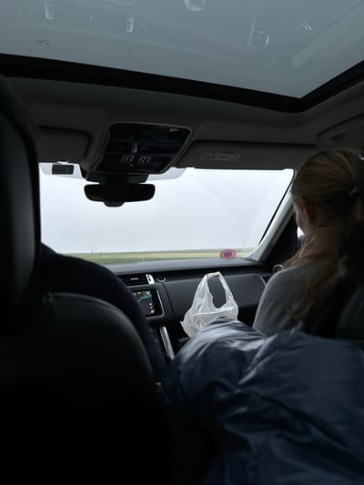 The image displays the interior of a modern vehicle, captured from a low, tilted perspective looking towards the front passenger side. The scene is rotated clockwise, positioning the bright white exterior background on the right side of the frame, creating a stark contrast with the darker car interior.

A person with light-colored hair (blonde or light brown) is visible in the upper left quadrant, leaning into the car. Their head and upper back are seen from above. On the right side of the frame, another person, wearing a dark blue jacket, is seated in what appears to be the front passenger seat. Their right arm is extended towards the center of the vehicle, holding a clear, crinkled plastic bag.

The car's dark interior features a dashboard and a center console with an infotainment screen displaying what looks like a navigation map with green lines. Air vents are visible on the dashboard. A light-colored sun visor is prominent on the left, bearing an "AIRBAG" warning label. Below the visor, a control panel with multiple buttons and a black circular knob is mounted. Dark car seats are present in the foreground and supporting the passenger. A small red circular sticker with a white symbol is affixed to the door frame or pillar near the open area.

The activity taking place is an exchange or interaction between the person leaning into the car and the passenger holding the plastic bag, possibly indicating a drive-through service, delivery, or a casual handover of an item.

Notable details include the bright, diffused light from outside, suggesting daytime, likely overcast conditions. A faint horizontal green line in the distant background hints at an outdoor, possibly rural or suburban setting. The visible text includes "AIRBAG" on the sun visor. The location context is Étréchy, France.
