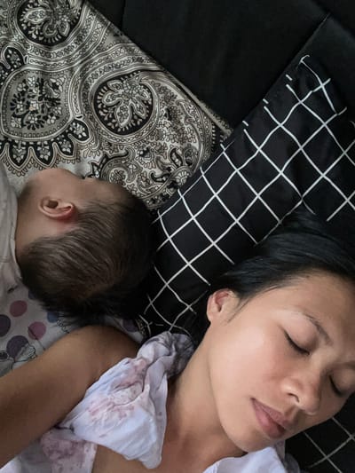 An overhead shot captures a serene moment of a woman and a baby sleeping together, likely co-sleeping or napping. The woman occupies the right side of the frame, lying peacefully on her back with closed eyes and a relaxed expression. Her dark hair is spread across a black pillow featuring a distinctive white grid pattern. She wears a light-colored top, possibly white or light pink, with subtle patterning.

To the bottom left, the baby's head is visible from the back, indicating it is sleeping on its side. The baby also has dark hair and appears to be wearing light clothing. They are both nestled amongst various bedding. The baby rests on a sheet or blanket adorned with an intricate black, grey, and white paisley-like design. Another lighter-colored sheet, possibly white with faint pink or purple polka dots or cartoon characters, is visible near the woman's chest and arm. The overall atmosphere is one of calm and intimacy, suggesting a peaceful resting period indoors. No discernible text is visible within the image. The location is specified as Ngasinan, Indonesia.