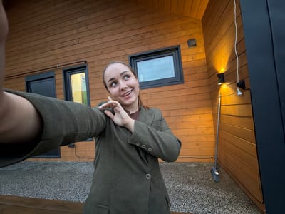 A young woman is taking a selfie outdoors in front of a wooden house. She is wearing a dark green blazer over a brown shirt. Her hair is pulled back in a ponytail. The house has dark trim around the windows and doors. There is a light fixture mounted on the wall and a broom leaning against the side of the house. The ground is covered in a grey carpet-like material. The woman is smiling and looking up and to her right. The image appears to be taken during the evening, as indicated by the artificial lighting. There is no information in the image to suggest this location is in Kamyennoye, Russia.