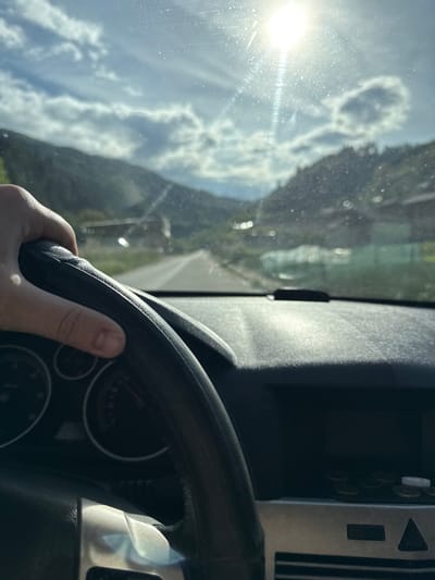 A driver's hand grips a black steering wheel inside a vehicle. Behind the wheel, through the windshield, is a landscape of tree-covered mountains and a partly cloudy sky, the sun a bright spot in the upper left corner. Based on the geography visible, it is highly probable that the scene is from Byala Reka, Bulgaria, given the mountainous terrain. The vehicle's dashboard and controls are visible to the right of the steering wheel.