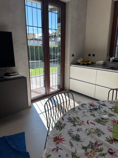 The image depicts an indoor scene, likely a kitchen and dining area, in what appears to be a modern home in Lissone, Italy. The room is brightly lit by natural light, suggesting it is daytime with clear skies visible through the large window.

On the left side of the frame, a large window with dark brown frames looks out onto a sunny exterior. Security bars are visible on the outside of the window, beyond which there is green foliage, possibly a hedge or grass, and a paved area. The blue sky and clouds are reflected in the windowpanes.

In the bottom left, a dark flat-screen television is mounted or placed against a textured gray wall. To its right, another dark piece of furniture, possibly a cabinet or dresser, is visible. A crumpled blue cloth or towel lies partially on the floor in the bottom right corner.

On the right side of the image, a kitchen area features white, handle-less cabinets with a sleek, minimalist design. A countertop with a fruit basket and some small items is visible. Above the countertop, two black wall sockets are mounted.

Dominating the upper right portion of the image is a round dining table covered with a vibrant floral tablecloth. The tablecloth features a pattern of red chili peppers, green herbs like rosemary and basil, and small purple and yellow flowers. Resting on the tablecloth are a pair of green-framed reading glasses, a white phone charger connected to a cable, and a small red roll of tape. A metal chair with a curved, slatted back is positioned next to the table. Bright sunlight streams through the window, casting distinct shadows on the floor and walls, particularly noticeable in the lower right portion of the scene. No discernible text is visible in the image.