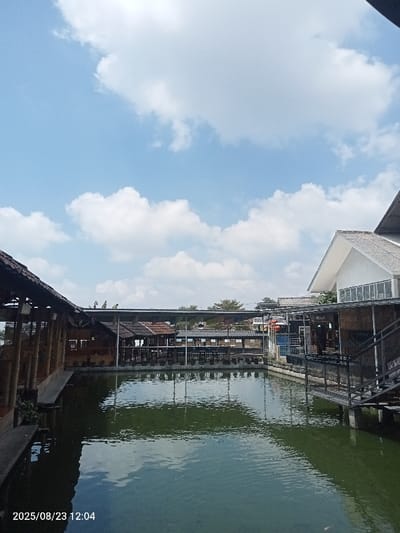 This image depicts an outdoor dining or recreation area in Palasari, Indonesia, captured at 12:04 PM on August 23, 2025. The scene is dominated by a large, dark green body of water occupying the lower half of the frame, reflecting the bright blue sky and white cumulus clouds overhead. On the left, a long, open-sided structure with a dark, possibly thatched or natural fiber roof is visible, supported by numerous light-colored vertical posts. Within this structure, rows of tables and chairs are arranged. In the background, across the water, additional similar structures with varying roof types, including corrugated metal, house more dining areas with tables and chairs. To the right, a more modern, two-story building features a white or light grey gabled roof and large windows on its upper level. Below this building, a dark metal staircase with railings descends, connecting to a lower, covered section that extends towards the water. No individuals are visible in the scene. The overall environment appears to be an unoccupied restaurant or resort situated around a pond or artificial lake under clear, sunny conditions.