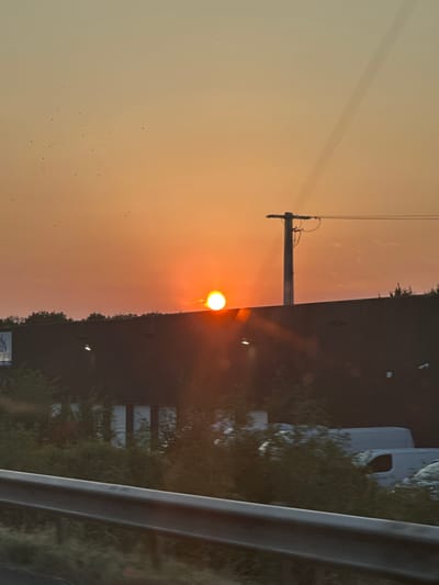 The image shows a sunset over a dark, flat-topped building, likely a warehouse or industrial facility, in Gondreville, France. The sun, a bright orange orb, is partially obscured by the top edge of the building and a utility pole with overhead wires. The sky is a gradient of orange and yellow, with some faint contrails visible. Numerous small birds are scattered across the sky. In the foreground, there is a dark guardrail, suggesting the photo was taken from a moving vehicle on a road. Behind the building, trees and parked white vans are visible. The overall scene depicts a commercial or industrial area at dusk.