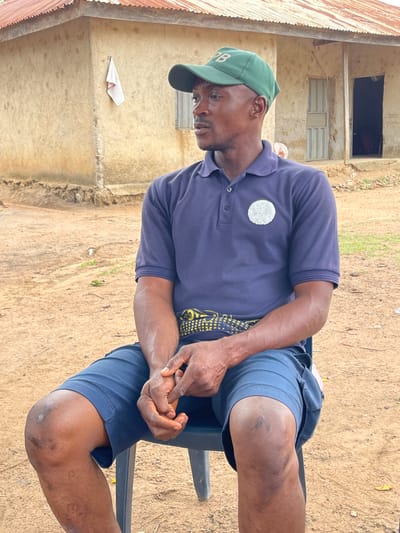 An outdoor, eye-level shot captures a man seated on a chair in a rural or semi-rural setting in Tabu, Nigeria. The man, with dark skin, is positioned in the foreground, looking downwards or to his left with a neutral expression. He wears a dark green baseball cap with a white letter "B" on the front, a dark blue short-sleeved polo shirt featuring a circular white, possibly textured, patch on the left chest, and dark blue shorts. His hands are clasped together in his lap, resting. Visible veins on his forearms and scuff marks on his knees suggest a history of physical activity or wear. A vibrant yellow and black patterned fabric is visible beneath his polo shirt, likely a wrapper or another layer of clothing.
The background consists of a simple, weathered building with light-colored, possibly painted, walls showing signs of age. An open, dark doorway and a window with horizontal metal slats are visible on the building. To the far left, a section of a corrugated metal roof is present. The ground beneath and around the man is dry, sandy earth. The scene is illuminated by bright daylight, indicating daytime and clear weather conditions. The overall atmosphere suggests an everyday moment in a rustic environment.
