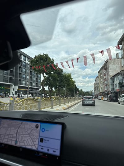 The image captures a street scene from inside a car in Edirne Merkez, Turkey. A road lined with trees and Turkish flags runs down the center of the frame. Several buildings and cars are visible on both sides of the street. The foreground includes parts of the car's interior, such as the dashboard, windshield, and infotainment screen displaying a map and music player.