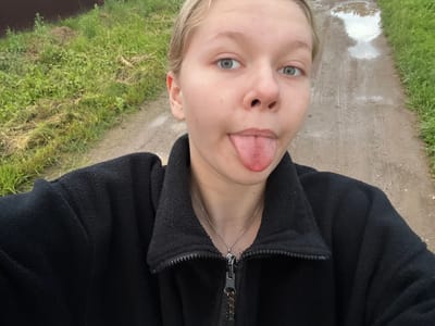 A young person with fair skin and light blue eyes is pictured sticking out their tongue, looking towards the camera. They have blonde hair and are wearing a black zip-up jacket and a delicate silver necklace. The image appears to be a selfie, taken outdoors. The background shows a wet, muddy path with tire tracks, bordered by lush green grass. A puddle reflects the sky. The lighting suggests it could be daytime, possibly after rain. The overall mood is playful and casual.