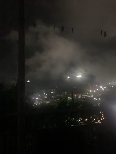 The image depicts a hazy, nighttime view of the city of Chiatura, Georgia. In the foreground, a dark, vertical structure, likely a pole or tree trunk, partially obscures the scene. Above, a wire is strung across the frame, with several dark, indistinct shapes hanging from it, possibly laundry or other objects.

The midground is dominated by an ethereal glow of lights from the city below, diffused by heavy fog or smoke. These lights are clustered and appear as soft, blurred orbs of white, yellow, and orange. A brighter, elongated light source is visible in the upper center of the city, possibly a stadium light or a powerful beacon. In the distance, the landscape slopes upwards, suggesting the hilly terrain of Chiatura. The overall impression is one of a dimly lit, somewhat mysterious urban environment shrouded in atmospheric conditions.