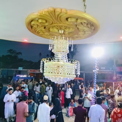 The image depicts a lively evening gathering under an ornate golden chandelier in Charfasson, Bangladesh. A large, multi-tiered chandelier adorned with crystal-like elements hangs from a decorative golden ceiling fixture, which also features subtle red lights.

A diverse crowd fills the area, consisting of men, women, and children. Many men are dressed in traditional kurtas or panjabis, while several women are wearing hijabs. People are standing, mingling, and observing their surroundings. Some individuals are seen looking at or holding mobile phones.

To the right, a bright light source on a pole wrapped with blue and white striped material illuminates a section of the crowd. The background shows a dark evening sky with silhouettes of trees and glimpses of dimly lit market stalls or structures, suggesting an outdoor public space, likely a market or an event venue. The scene conveys a communal and active atmosphere during what appears to be a social or festive occasion at night. No discernible text is visible in the image.