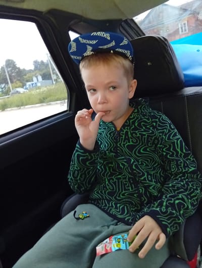 A young Caucasian boy with blonde hair, wearing a blue baseball cap and a green and black patterned hoodie, sits in the back seat of a car. The cap has the word "KNICKS" written on it. His right hand is near his mouth, and he is looking towards the camera. He is wearing a green t-shirt or hoodie underneath the patterned one and green pants with a small toy attached. The car appears to be parked or moving slowly on a street in Novoilinsky, Russia, with trees and buildings visible through the window. The interior of the car is dark, with black upholstery.