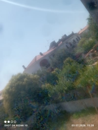 The image, taken from a slightly elevated angle, shows a view of a building with a red-tiled roof and multiple windows, partially obscured by lush green trees and foliage. The sky is bright blue and appears slightly hazy. The overall scene suggests an outdoor setting in a residential area. The bottom left corner of the image displays the text "SHOT ON REDMI 10", and the bottom right corner shows the date "03.07.2026" and time "15:28". The image has some visual artifacts, including circular distortions and color aberrations, particularly in the lower half. The scene does not provide specific details that can definitively place it in Ardea, Italy, beyond what might be inferred from general architectural styles or vegetation, which are not unique to that location.