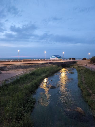 The image depicts a small, man-made stream flowing through a grassy area. The stream is illuminated by the reflection of lights from a nearby bridge and a small building labeled "L'AIRE A LA PLAGE." The building has a blue roof and white walls. Beyond the stream, a paved walkway runs parallel to the water, leading to a promenade with benches and lampposts. The promenade overlooks a body of water, likely the sea, with a distant horizon. The sky is cloudy and twilight, suggesting it is either early morning or late evening. The scene appears to be in a public park or recreational area in Veules-les-Roses, France. No people are clearly visible in the frame, though shadows suggest potential presence near the building. The overall atmosphere is peaceful and serene.