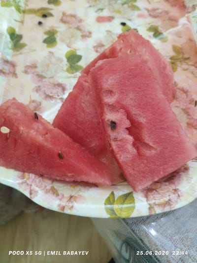 The image displays a close-up of three slices of watermelon arranged on a plate with a floral pattern. The watermelon slices are a vibrant pink and appear juicy, with some seeds visible. The plate has a light-colored background adorned with pink and white flowers and green leaves, giving it a decorative appearance. The setting appears to be indoors, possibly a table, given the textured surface visible below the plate.

The bottom left corner of the image contains visible text: "POCO X5 5G | EMIL BABAYEV". In the bottom right corner, there is a timestamp: "25.06.2026 22:44". This indicates the image was taken on June 25, 2026, at 10:44 PM, suggesting it was taken in the evening. The location context provided is Sabail Raion, Azerbaijan. There are no people or animals visible in the image. The focus is solely on the watermelon and the plate.