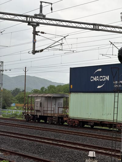 This image depicts a freight train in Saphale, India, moving along a railway track beneath an overhead electrical catenary system. The train consists of multiple wagons. The foremost wagon visible is a grey, open-topped carriage with a visible serial number "871222 14843" and the designation "ECoR". It appears to be carrying a stacked load. On top of this wagon, and extending further back, are two large shipping containers. The upper container is dark blue and prominently displays the logo and text "CMA CGM". The container below it is a pale green color. The railway tracks are laid on a bed of dark ballast. In the background, a lush green, mountainous landscape with trees and foliage is visible under an overcast sky. A metal pole with electrical insulators and wires is prominent in the middle ground, supporting the catenary system. Another metal pole, part of a signal or utility structure, is visible on the right side of the frame.