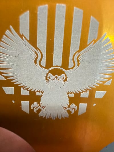 The image shows a close-up of an etched eagle design on an orange surface. The eagle is depicted with spread wings, facing forward, with its talons extended. Behind the eagle, there are vertical white stripes and a geometric pattern of white squares and rectangles. The surface appears to be a metallic or lacquered material, as evidenced by the reflections and the glossy finish. There is no indication of any specific location in the city of Grapevine, United States of America, or any people, actions, or interactions. The focus is solely on the etched design.