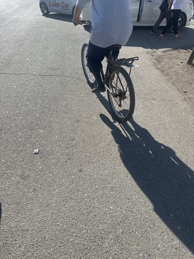 An external, eye-level, slightly tilted, medium shot shows a person on a bicycle, facing away from the camera. The person is wearing a grey t-shirt and dark pants. The bicycle is dark-colored with a rear rack. The scene appears to be a street in the city of Kara-Suu, Kyrgyzstan. To the left of the cyclist, a white car with the text "Lider Car detailing" is partially visible. Two individuals are walking behind the car. The ground is asphalt with visible cracks. A dark shadow of the cyclist and their bicycle is cast on the asphalt to the right. A small white object, possibly debris, is on the ground in the foreground. The lighting suggests it is daytime with direct sunlight.