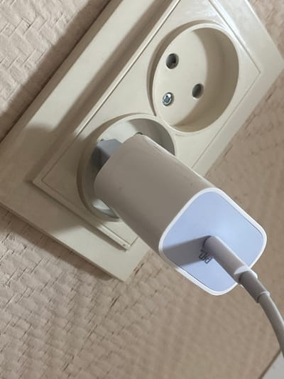 The image shows a close-up, high-angle shot of a power adapter plugged into a wall socket. The socket is a light beige color and has two round holes for prongs and a small screw in the center. The power adapter is also light beige with a light blue accent on the end where the cable connects. The cable is gray and snakes away from the adapter. The wall surrounding the socket is textured, resembling wallpaper or painted plaster, with a light brown or beige color. The lighting suggests an indoor environment. The image does not provide enough information to determine its location in the city of Афанасьево, Russia.