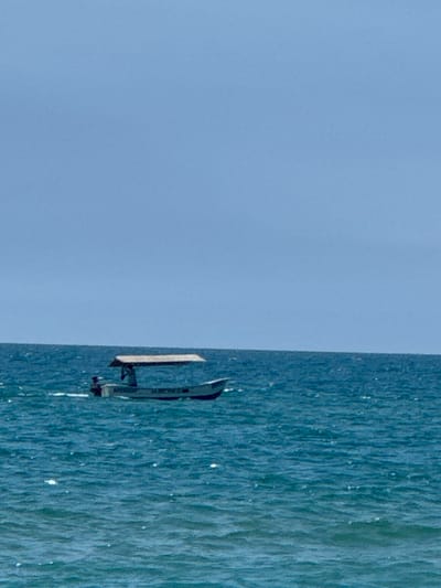 A low-angle, medium shot captures a small boat with a canopy sailing on the blue ocean under a clear, pale blue sky. The boat is positioned on the right side of the frame, moving from left to right. Two individuals appear to be on board the boat. The water exhibits choppy waves, with whitecaps reflecting the sunlight. The scene is set on the coast of Vityazevo, Russia, a resort town on the Black Sea, known for its sandy beaches and shallow waters.