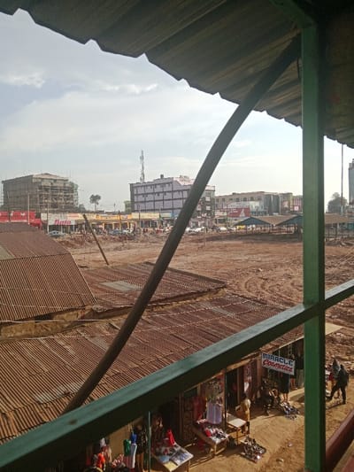 The view from a rooftop in Kapsabet, Kenya (latitude: 0.202, longitude: 35.098) shows a market area. The market is mostly empty except for a few people walking around. Some stores are visible on the right side of the image, with goods displayed on shelves. There is a large empty space in the center of the image, possibly a parking lot or a construction site. The buildings in the background appear to be residential and commercial. There are several trees in the distance. The sky is clear and blue with some clouds. The image is taken from a building under construction. The building has a corrugated metal roof. The railing of the building is green and the railing of the roof is white. The surrounding area is dusty.