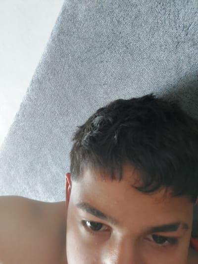 A high-angle, close-up image depicts the upper body and face of a young individual, situated in Glava, Bulgaria. The subject has short, dark, textured hair, dark eyebrows, and dark eyes looking downwards and slightly to the left. Their skin tone is light to medium, and a portion of their bare shoulder is visible on the left. The background is composed of a textured, mottled grey surface, possibly fabric or carpeting, covering the right and upper areas, meeting a plain white surface on the upper left along a diagonal line. The scene is illuminated by diffused lighting.